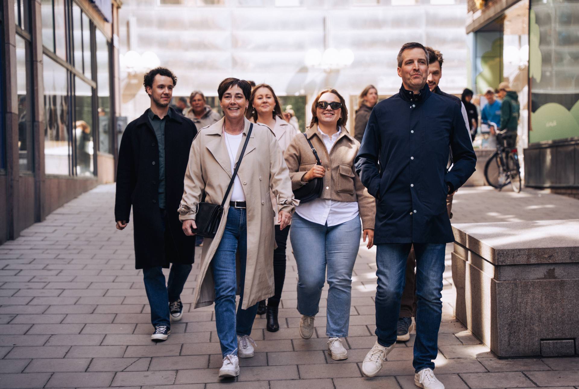 A group of people walk confidently down a city street, wearing casual jackets and jeans, with modern buildings in the background.