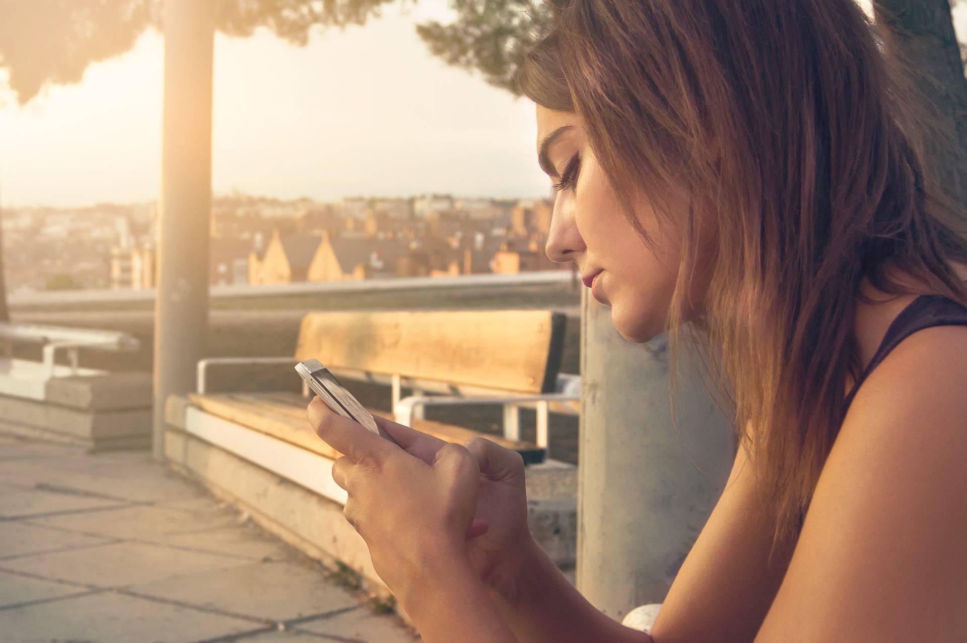 Kvinna som sitter utomhus på en bänk, tittar på sin smartphone, med en stadsbild i bakgrunden vid solnedgång.